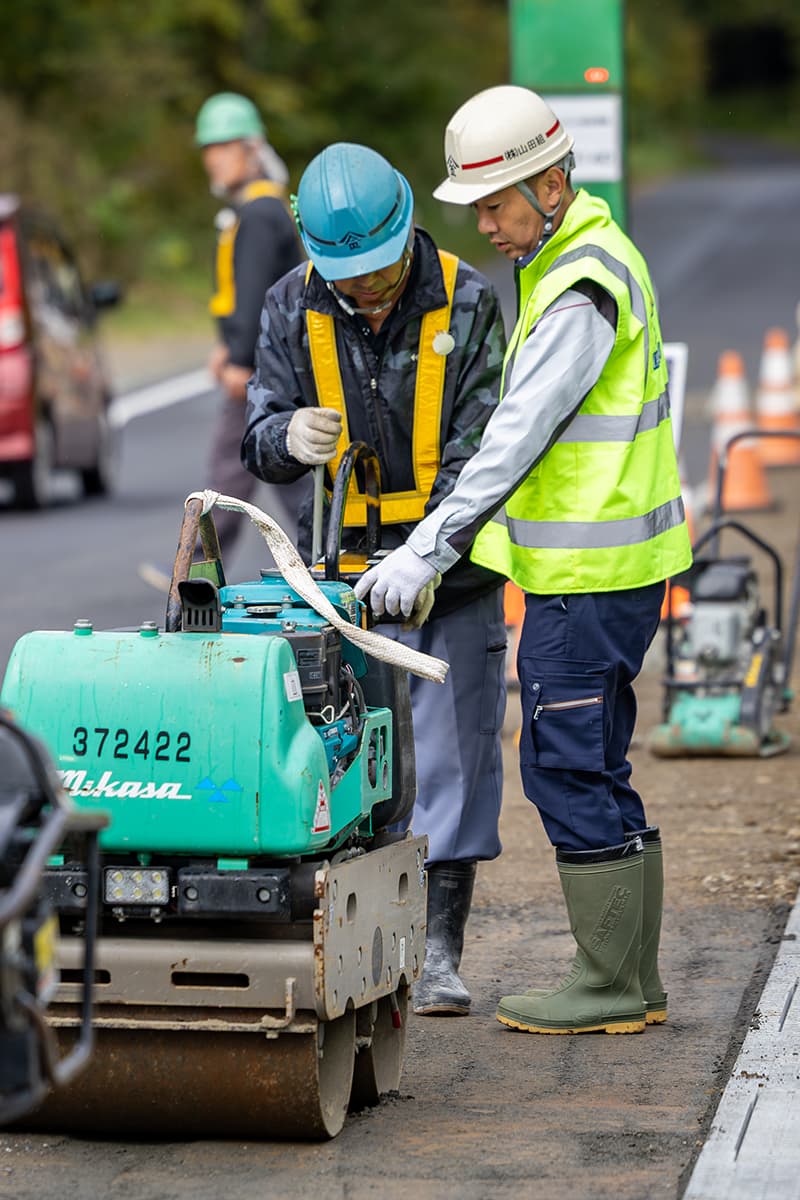 道路工事現場で白いヘルメットとオレンジ色の安全ベストを着た現場監督が、完成したばかりのアスファルト道路を背に立っている様子