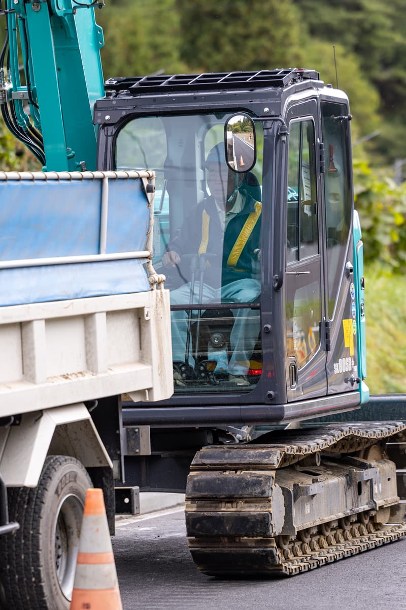 道路工事現場で白いヘルメットとオレンジ色の安全ベストを着た現場監督が、完成したばかりのアスファルト道路を背に立っている様子