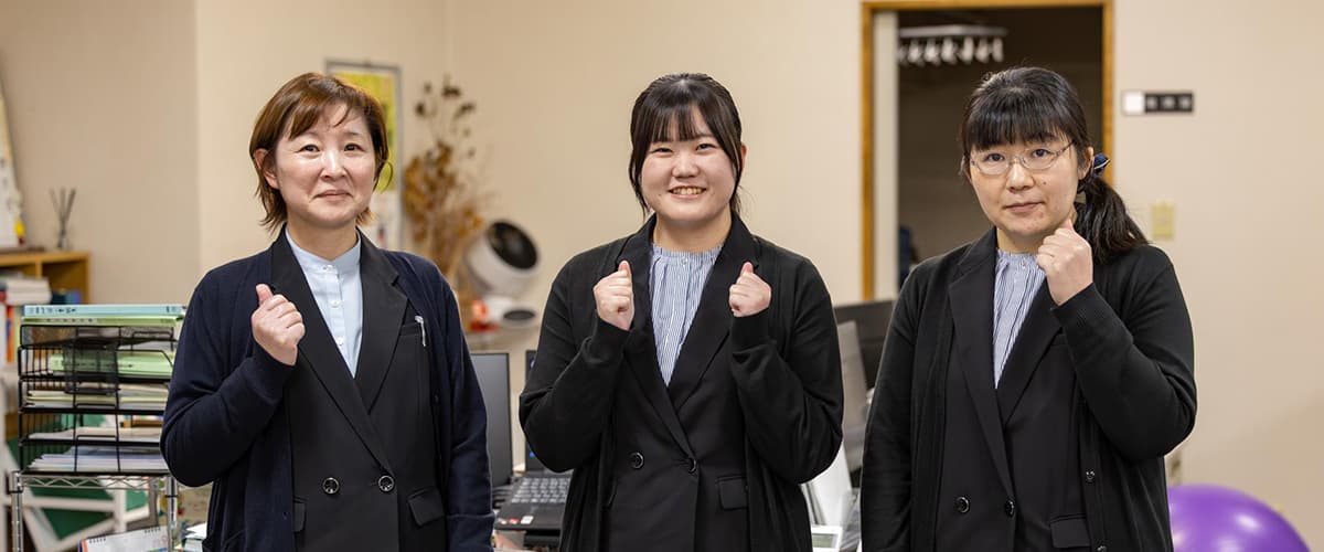 明るいオフィスで黒い作業服を着た3人の社員が、テーブルに向かって資料を見ながら笑顔で打ち合わせをしている様子
